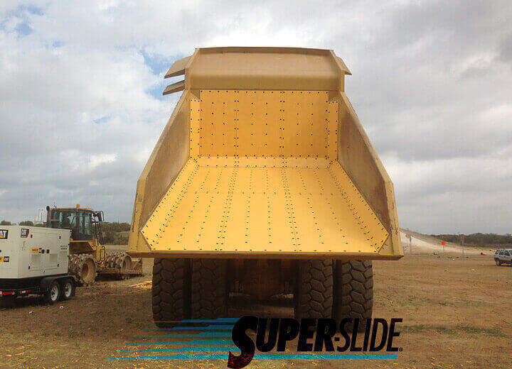 A large yellow dump truck with an empty bed features a Super-Slide Yellow Quartz UHMW Liner. Parked on dirt ground and viewed from the rear, it has construction equipment and a cloudy sky in the background. "SUPERSLIDE" appears below.